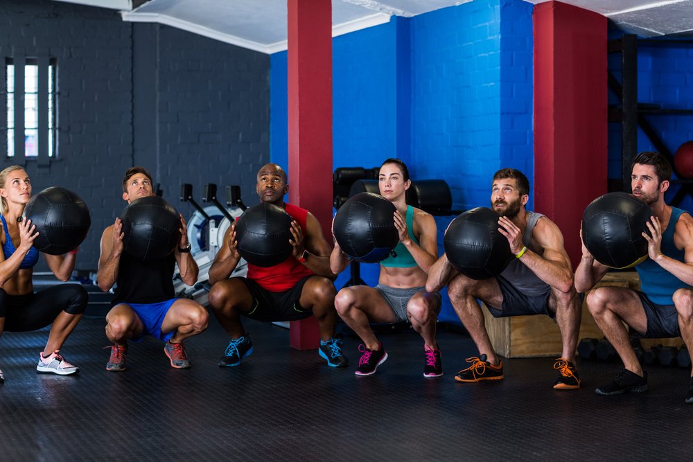 Friends holding exercise ball while crouching in gym-1