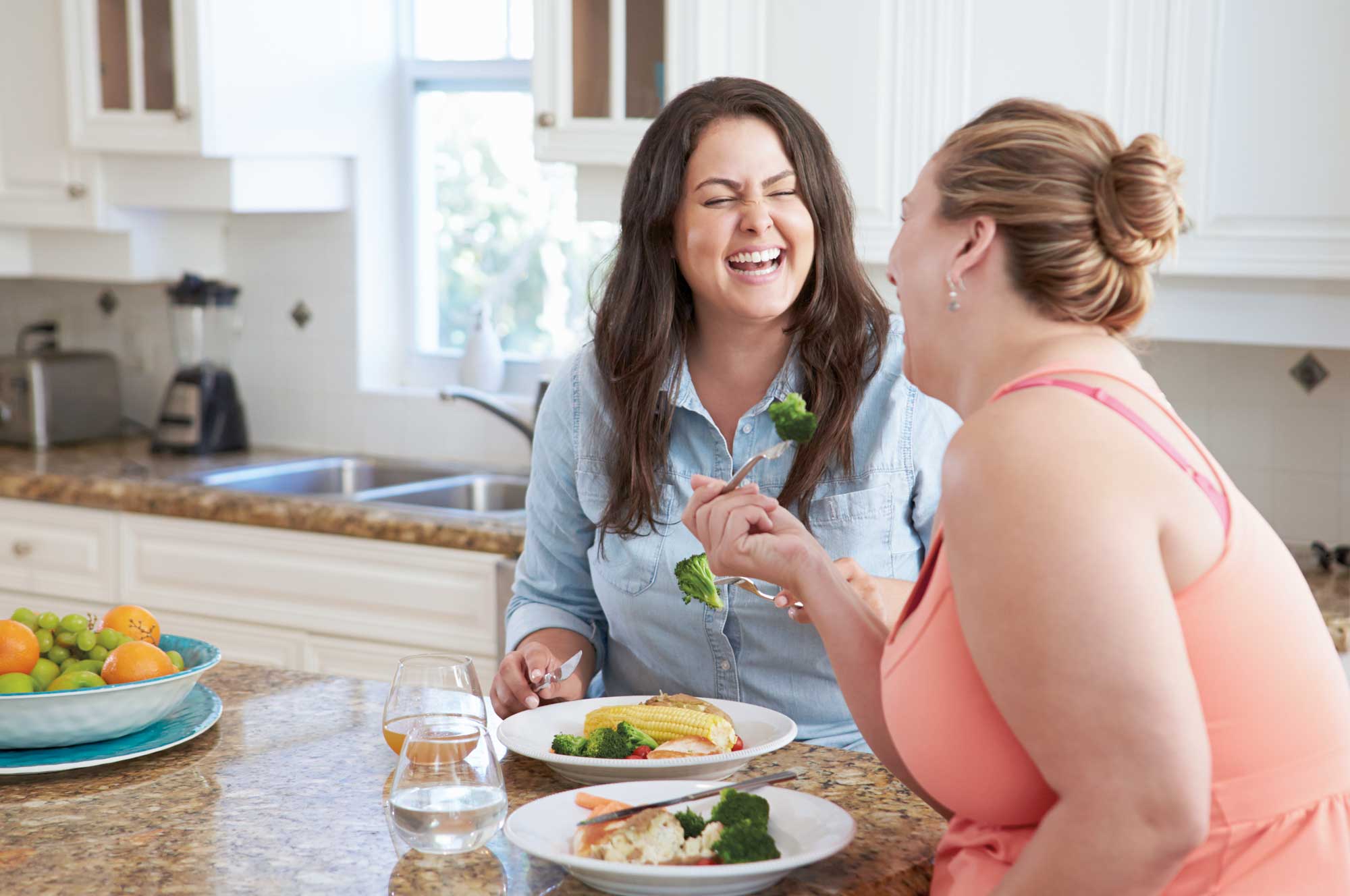 Zwei Frauen sitzen in einer Küche am Tisch, essen gemeinsam und lachen miteinander